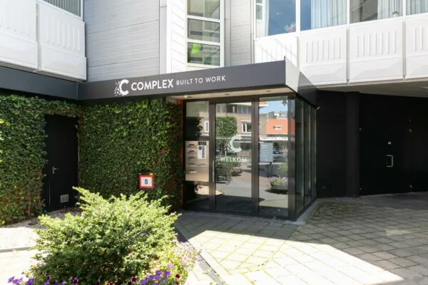 Ingang van het bedrijfsverzamelgebouw Complex aan de 1e Hogeweg met glasdeur, begroeiing en een B-ingangsaanduiding.